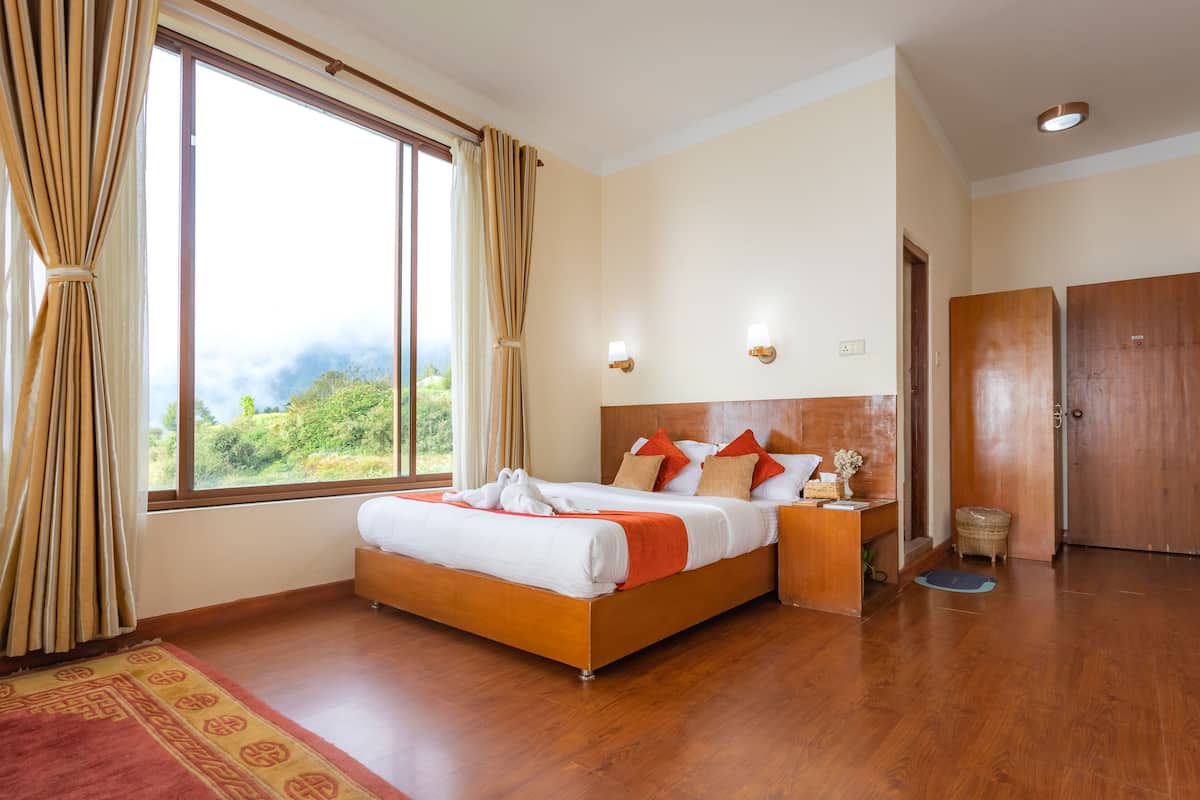 Una habitación de hotel con una cama grande, cabecero de madera, dos mesitas de noche y una ventana con cortinas que ofrece vistas a zonas verdes.