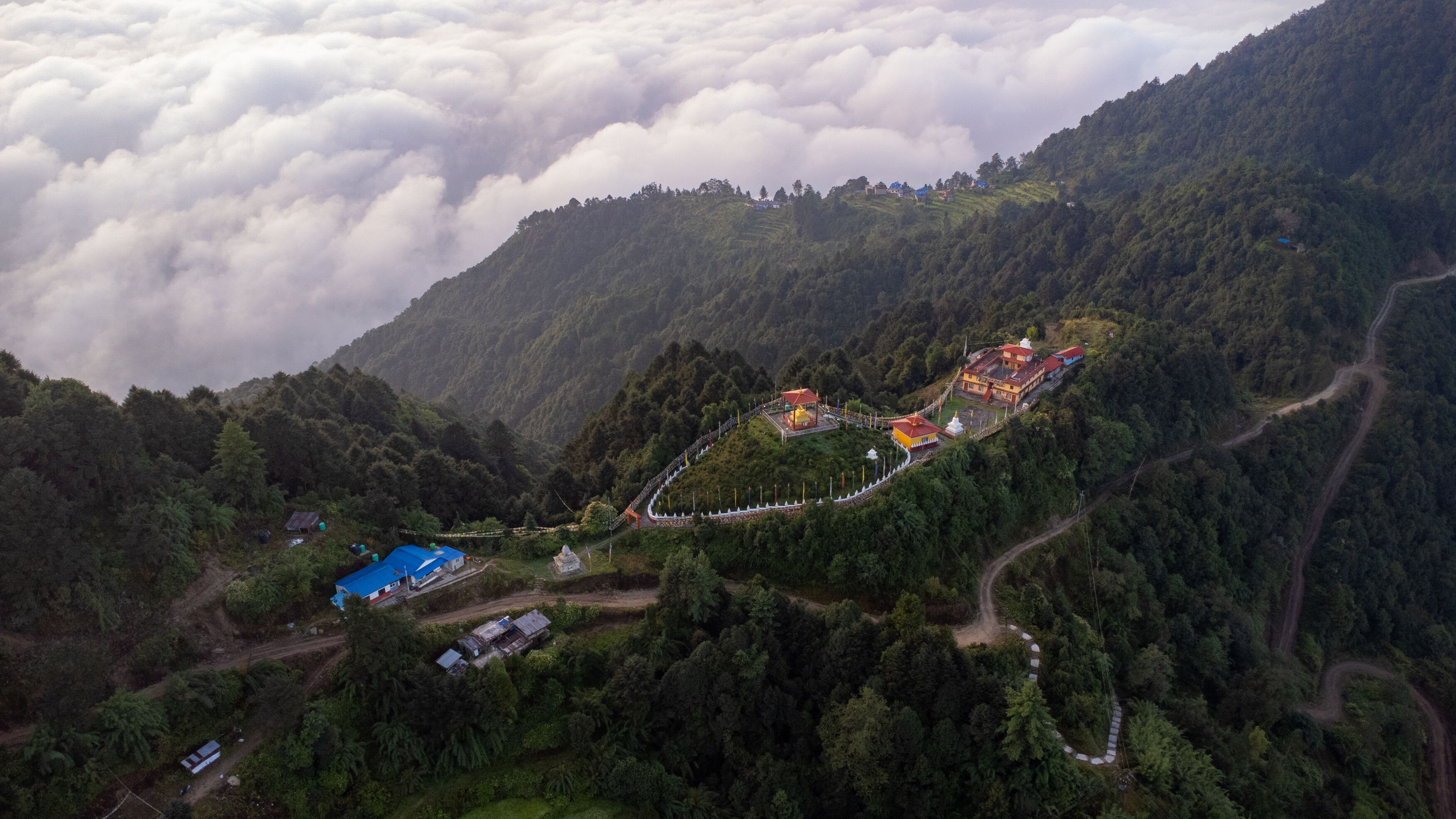 An aerial view of a mountainous area with a temple complex surrounded by lush greenery and a winding road.