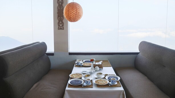 A dining area with a table set for two, a hanging light fixture, and a view of the sky through large windows.