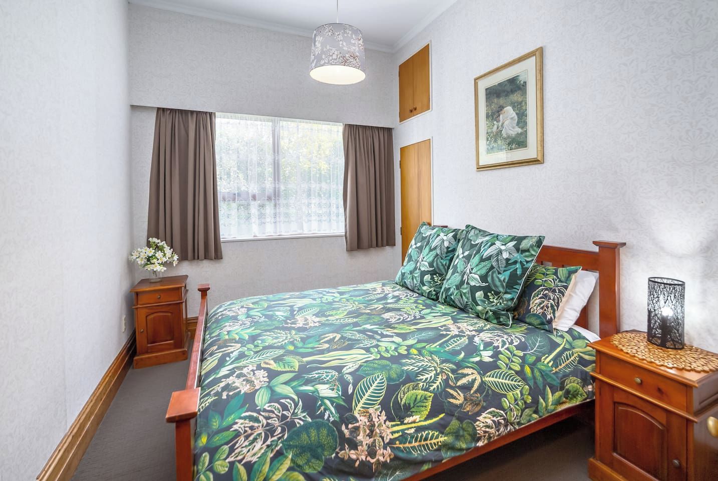 A bedroom with a bed, wooden nightstands, a hanging light fixture, a window with curtains, and a framed picture on the wall.