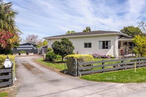 A single-story house with a paved driveway, a wooden fence, and a variety of shrubs and small trees.