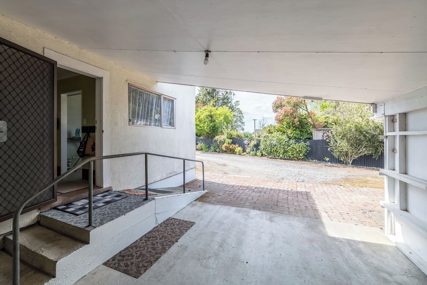 A covered outdoor area with a staircase leading to a door, a metal railing, and a view of a garden with trees and a fence.