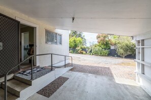 Un espacio exterior cubierto con una escalera que lleva a una puerta, una barandilla de metal y vistas a un jardín con árboles y una cerca.