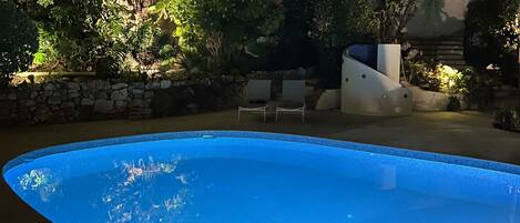 Una piscina rodeada de abundante vegetación y un edificio blanco con múltiples ventanas y balcones.
