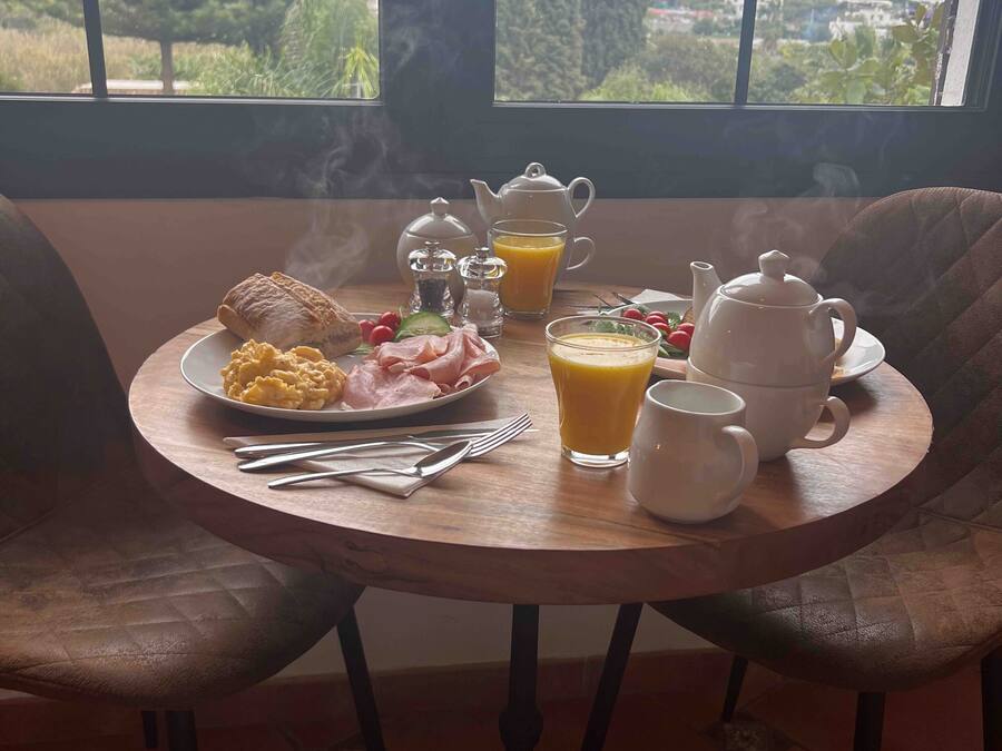 A breakfast table with eggs, ham, toast, and juice, set in a room with a view of trees and buildings through large windows.