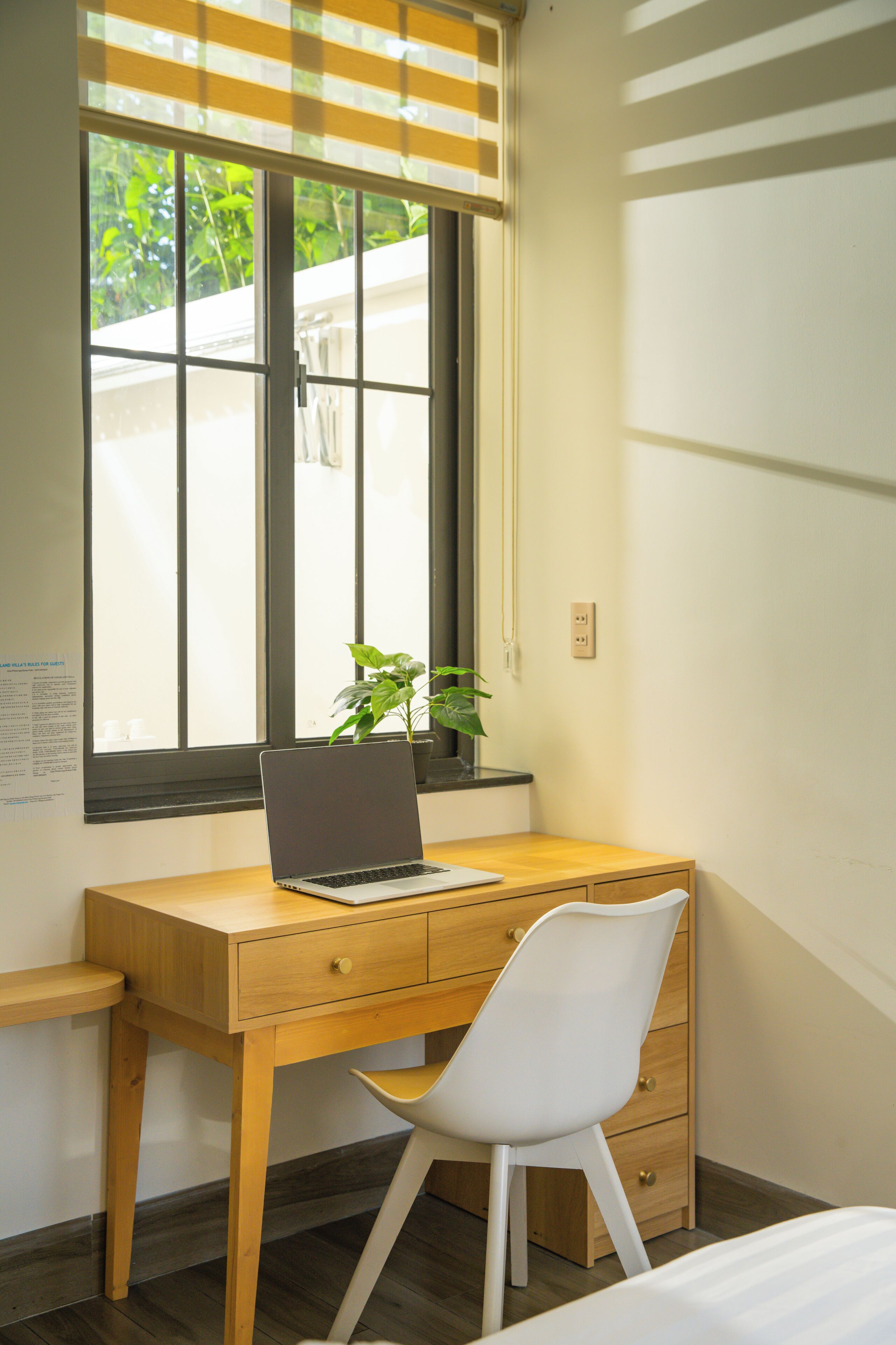 A wooden desk with a laptop and a potted plant, a white chair, and a window with blinds.