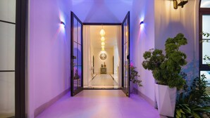 Un pasillo con una puerta de cristal que da a una habitación, una planta en maceta y una lámpara fijada a la pared.