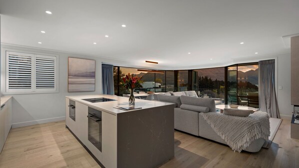 A modern kitchen with a central island, a dining area with a sofa, and a large sliding glass door leading to a scenic outdoor view. - NEW! Sunset Views | Queenstown Central | Lake & Mountain View (Queenstown)