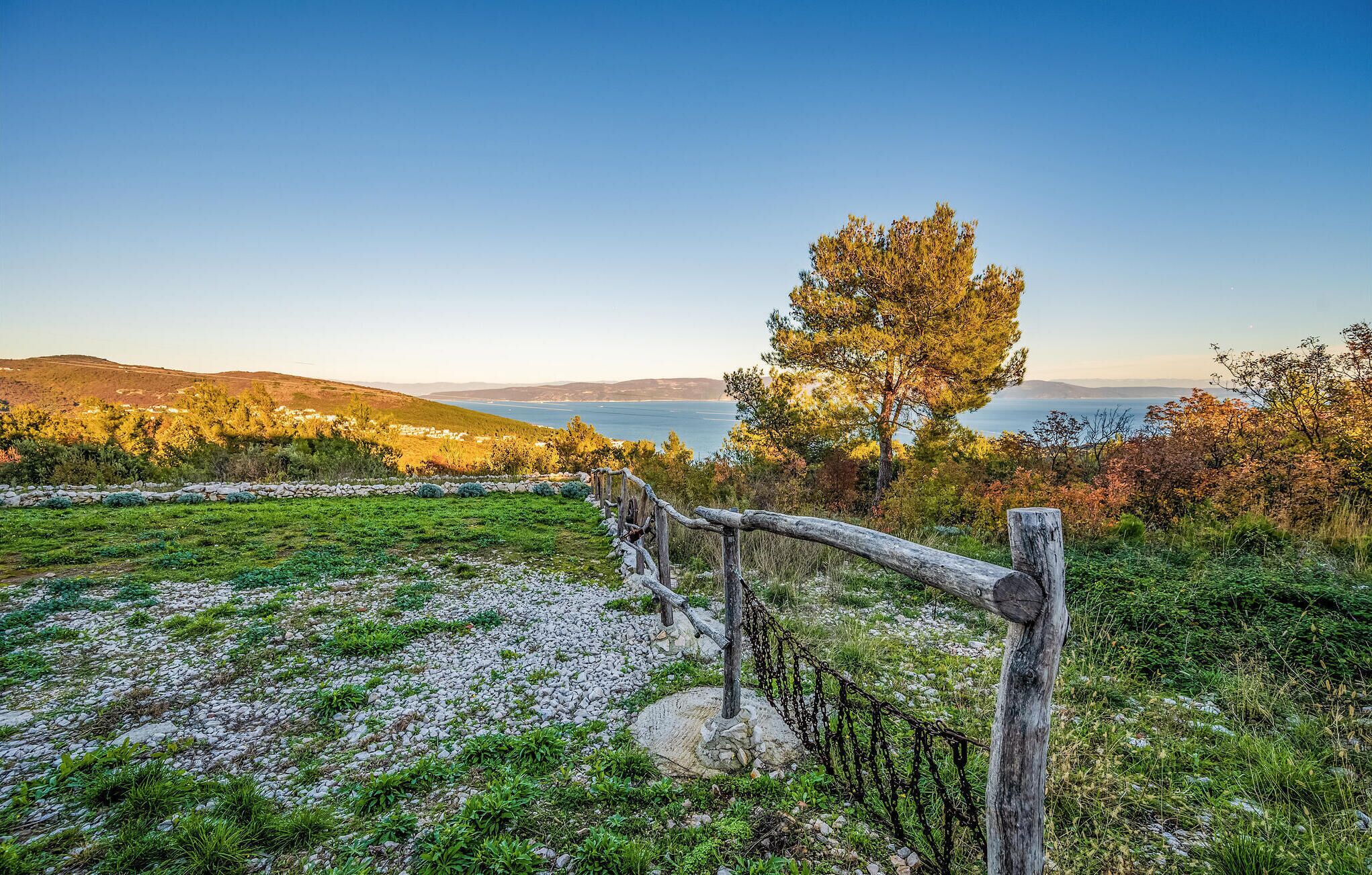 Un sentier de pierres mène à une clôture en bois rustique, d’où l’on peut admirer un paysage pittoresque offrant une vue dégagée sur la mer et les collines lointaines.