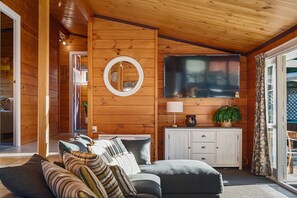 A cozy cabin interior with a wooden interior, a television mounted on the wall, a sofa with cushions, and a cabinet with drawers. - Highfield Ride - Spa Pool, Ride the Redwoods (Rotorua)