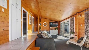 A modern log cabin interior with a large sofa, a striped armchair, a dining table, and a TV mounted on the wall. - Highfield Ride - Spa Pool, Ride the Redwoods (Rotorua)