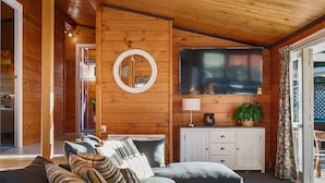 A cozy cabin interior with a wooden ceiling, a television mounted above a white cabinet, and a comfortable seating area with a sofa and pillows. - Highfield Ride - Spa Pool, Ride the Redwoods (Rotorua)