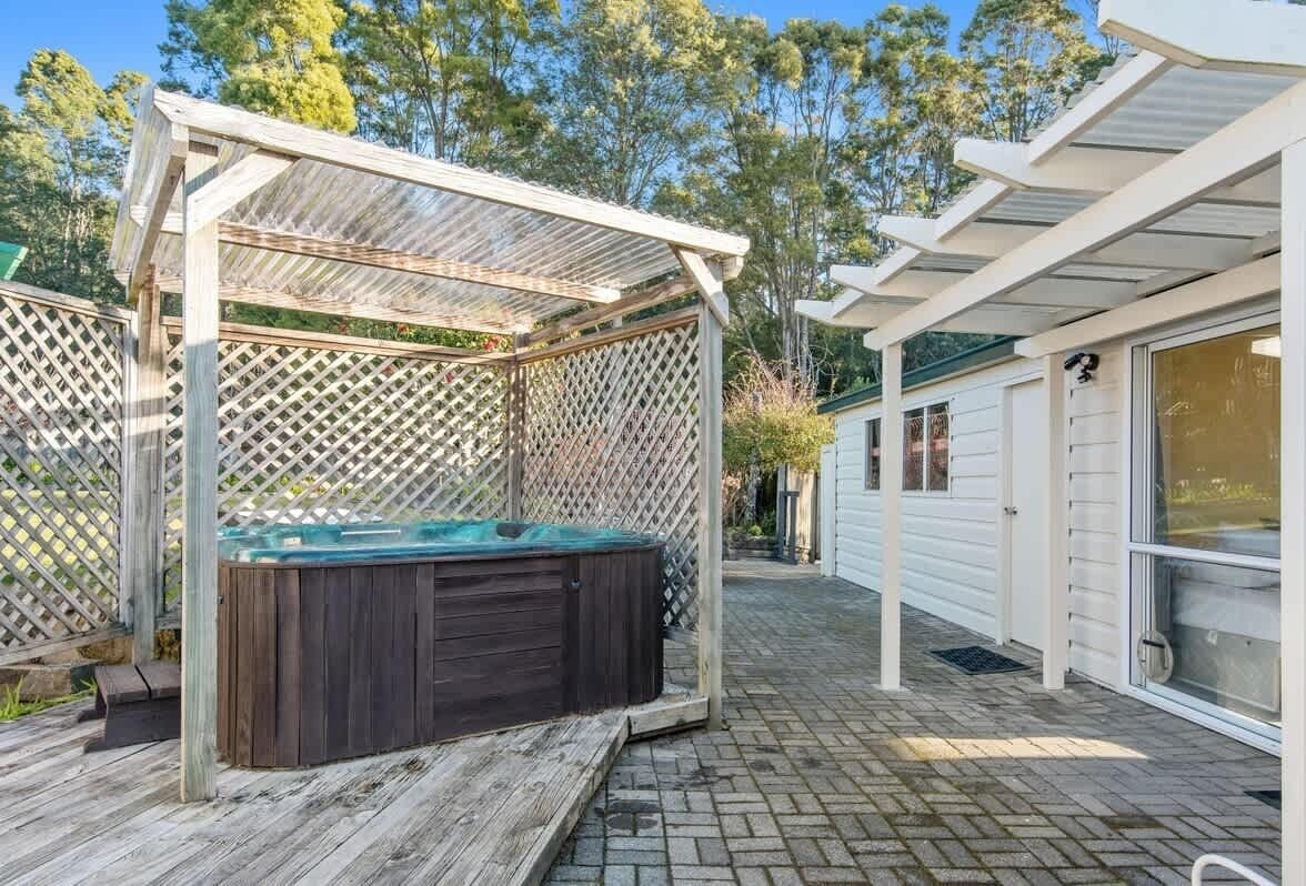 Un jacuzzi bajo una pérgola de madera con un cerramiento tipo celosía, un patio de ladrillo y un edificio blanco con un garaje al fondo.