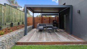 A modern outdoor patio with a wooden deck, a gray sofa, and a table. - The Sanctuary - Modern, Spa Pool, Sleeps 4 (Rotorua)