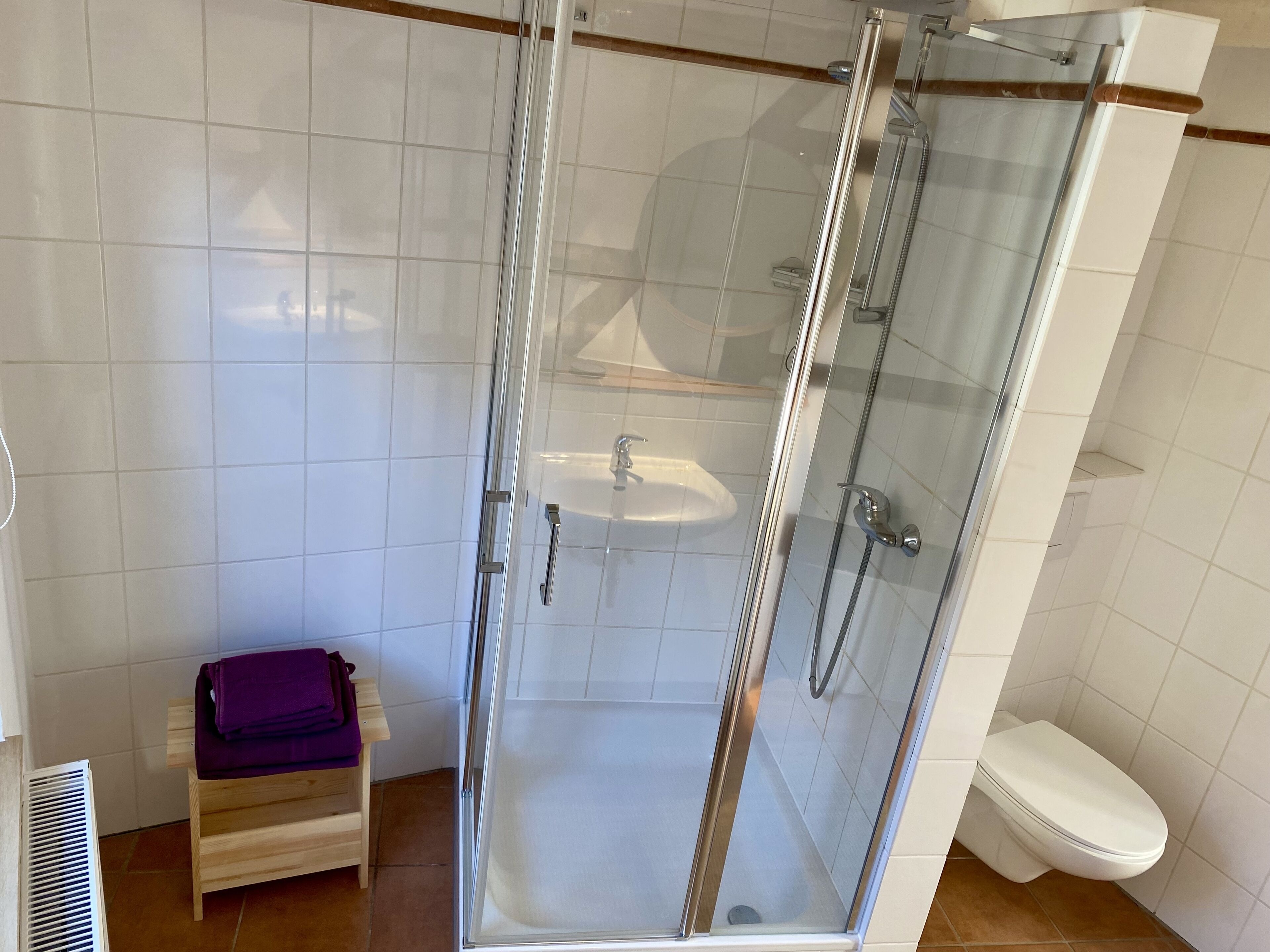 Une douche fermée par des parois de verre, avec un lavabo blanc et un tabouret en bois.