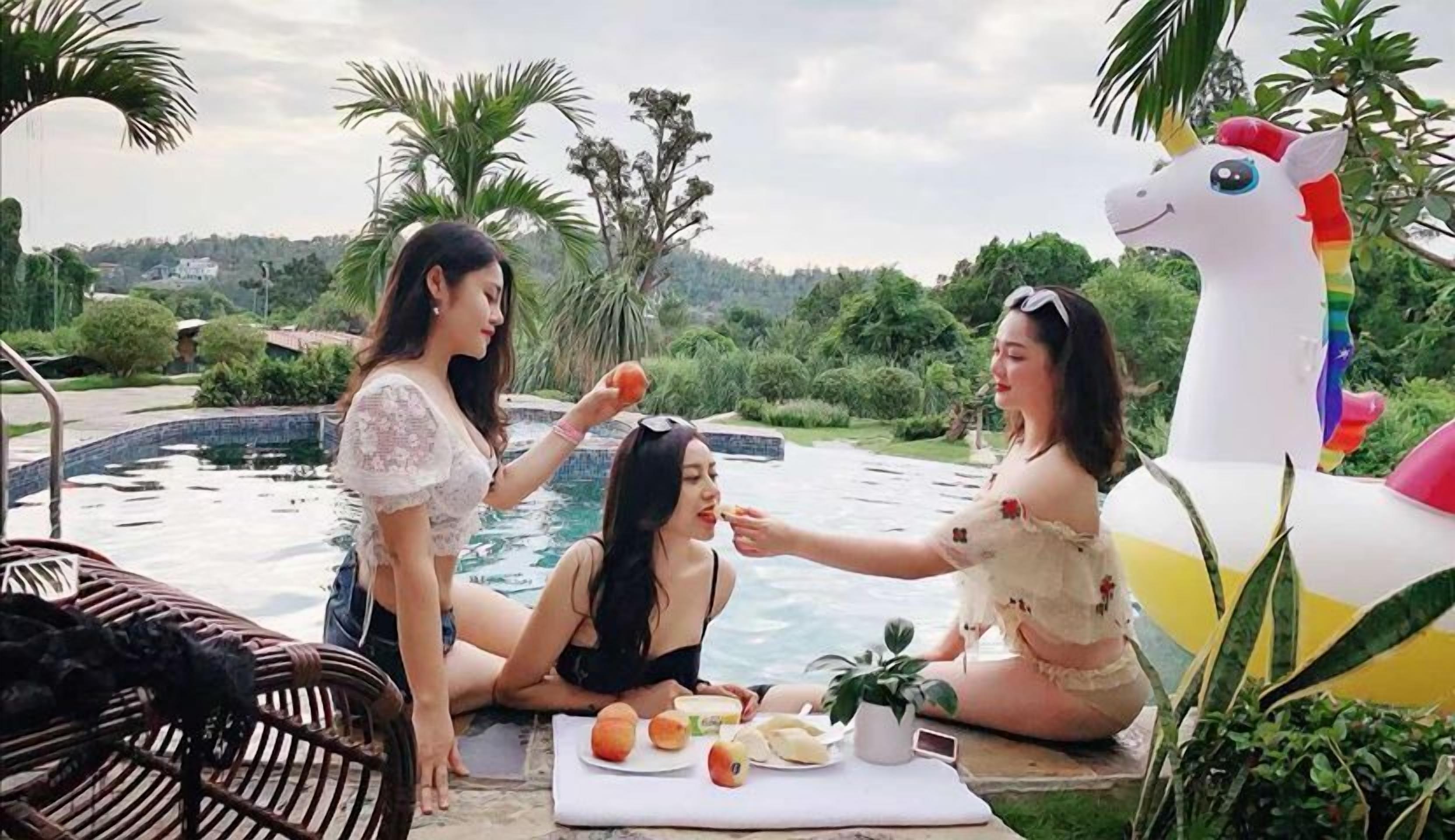 Three women by a pool with a unicorn float, surrounded by greenery and a view of hills.