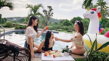 Three women by a pool with a unicorn float, surrounded by greenery and a view of hills.