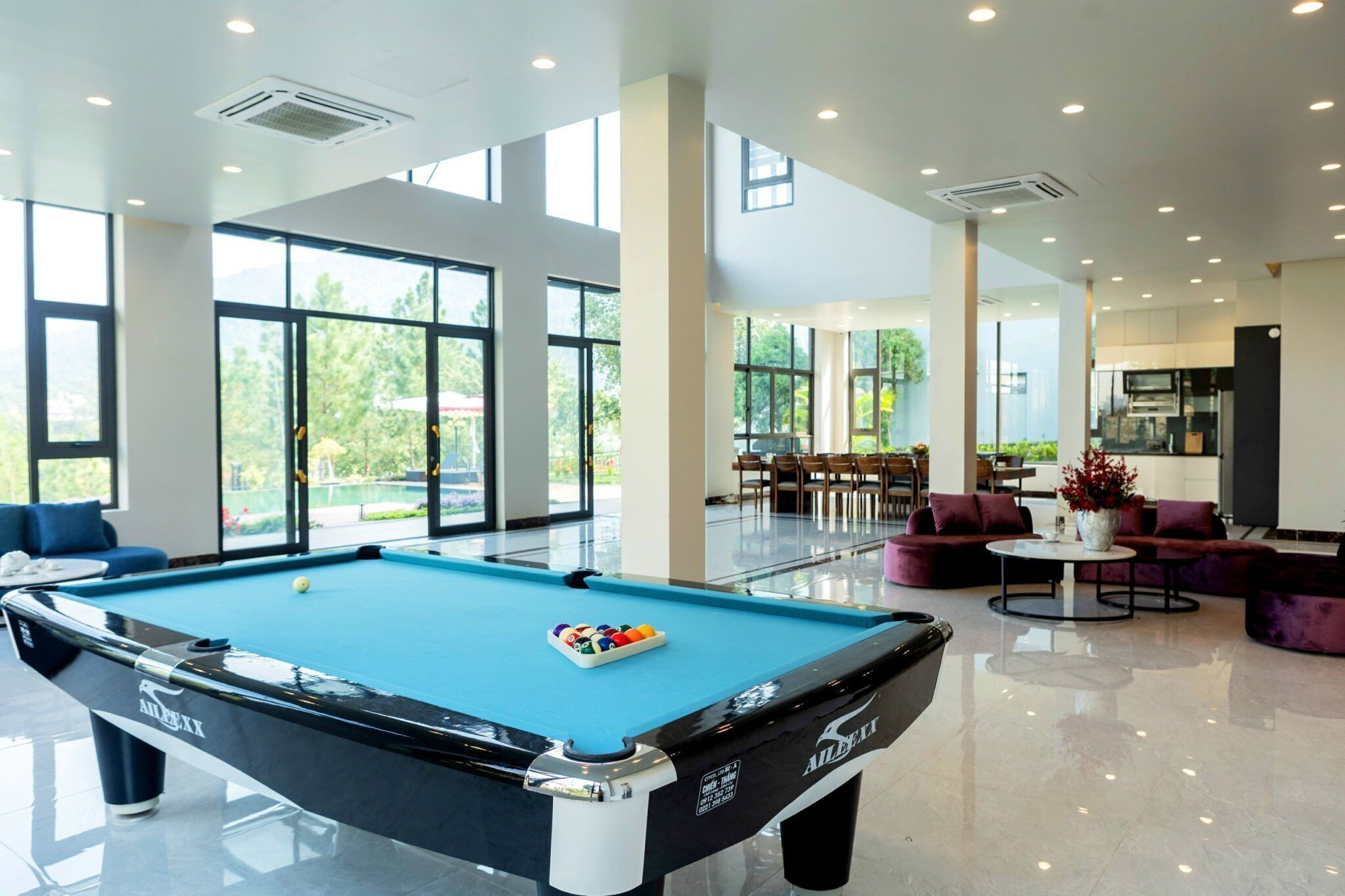 A pool table in a spacious room with large windows, a dining area, and a kitchen.