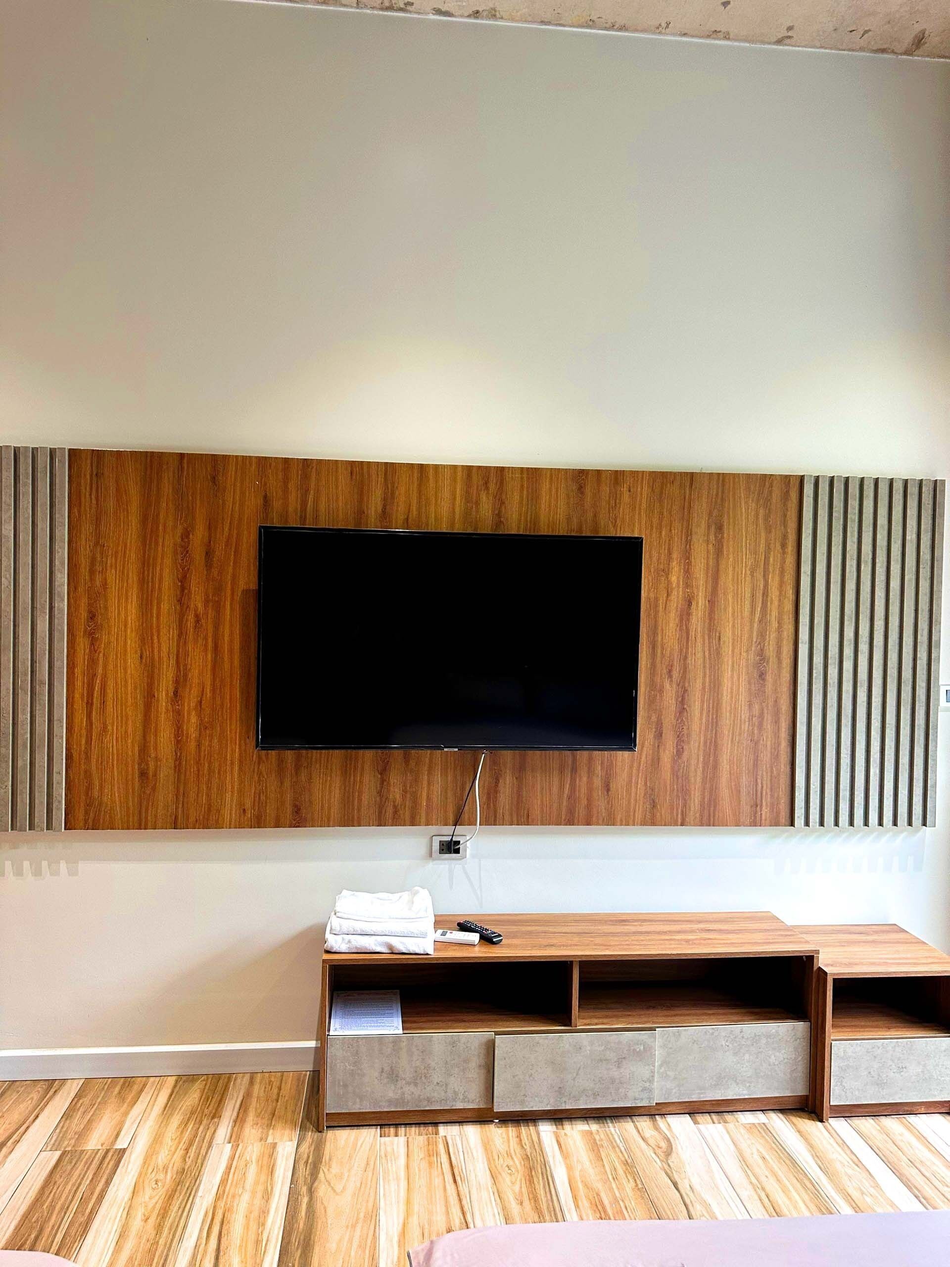 A modern living room with a flat-screen TV mounted on a wooden panel wall, a wooden entertainment unit with drawers, and a wooden floor.