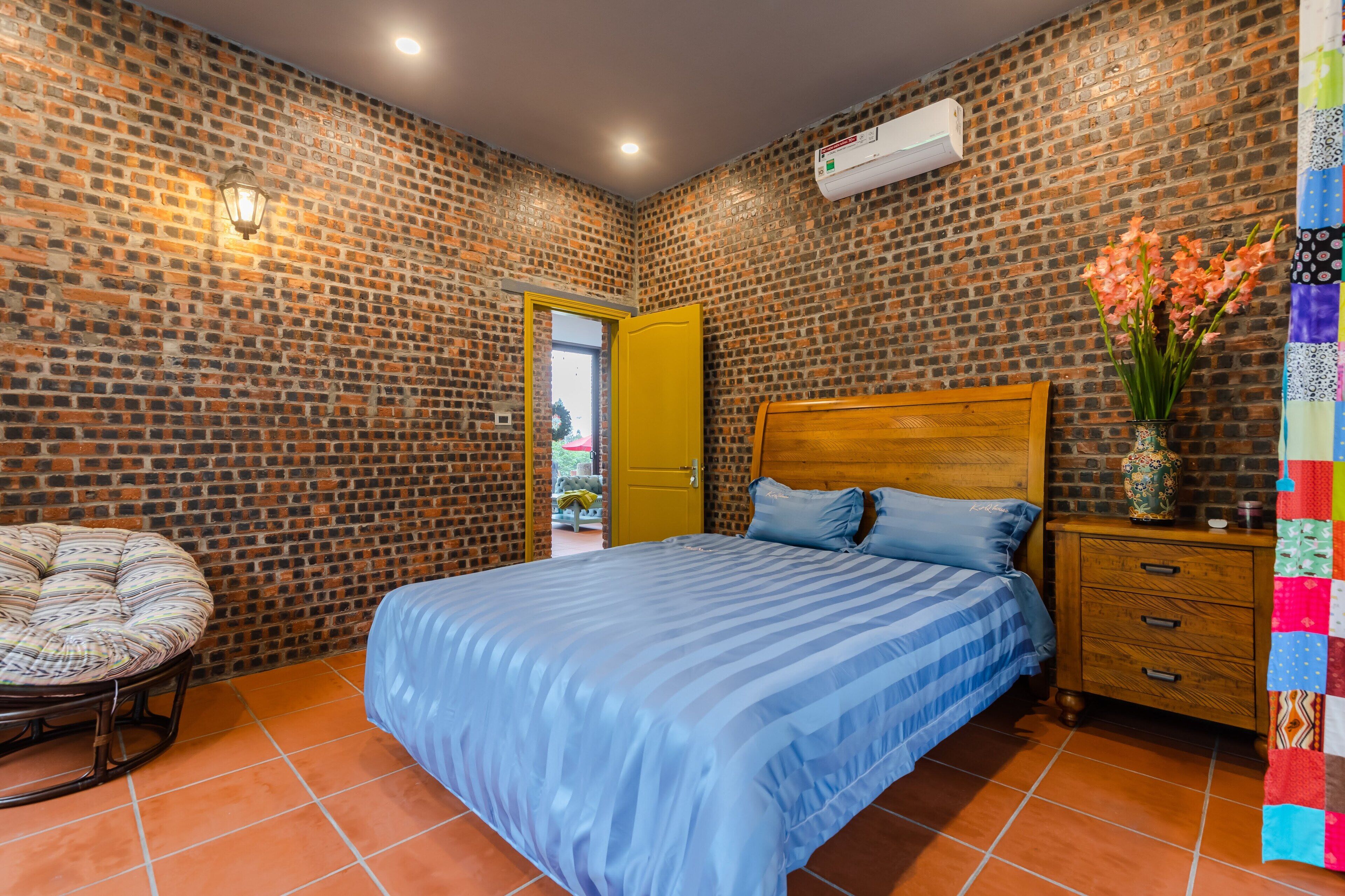 A bedroom with a brick-patterned wall, a wooden bed frame, a blue-striped bedspread, a wooden nightstand with drawers, and a wicker chair.