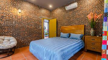 A bedroom with a brick-patterned wall, a wooden bed frame, a blue-striped bedspread, a wooden nightstand with drawers, and a wicker chair.