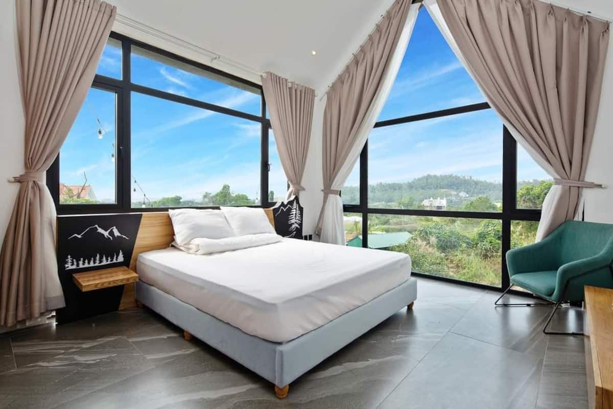 Ein modernes Schlafzimmer mit einem großen Bett, einem Nachttisch, einem grünen Sessel und Blick auf eine grüne Landschaft durch große Fenster.