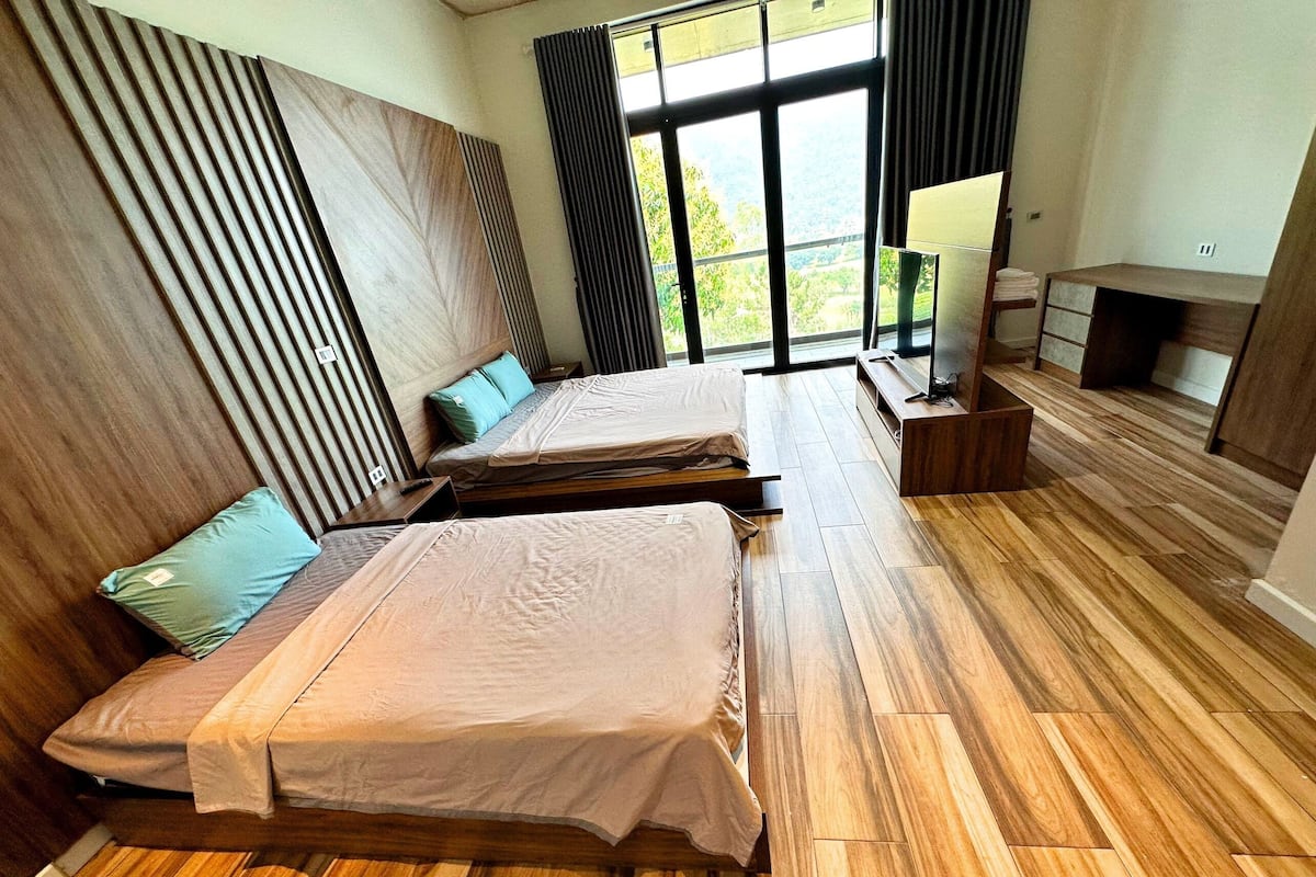 Ein modernes Hotelzimmer mit zwei Betten, Holzboden, einem großen Fenster mit Aussicht und einem kleinen Schreibtisch mit Lampe.