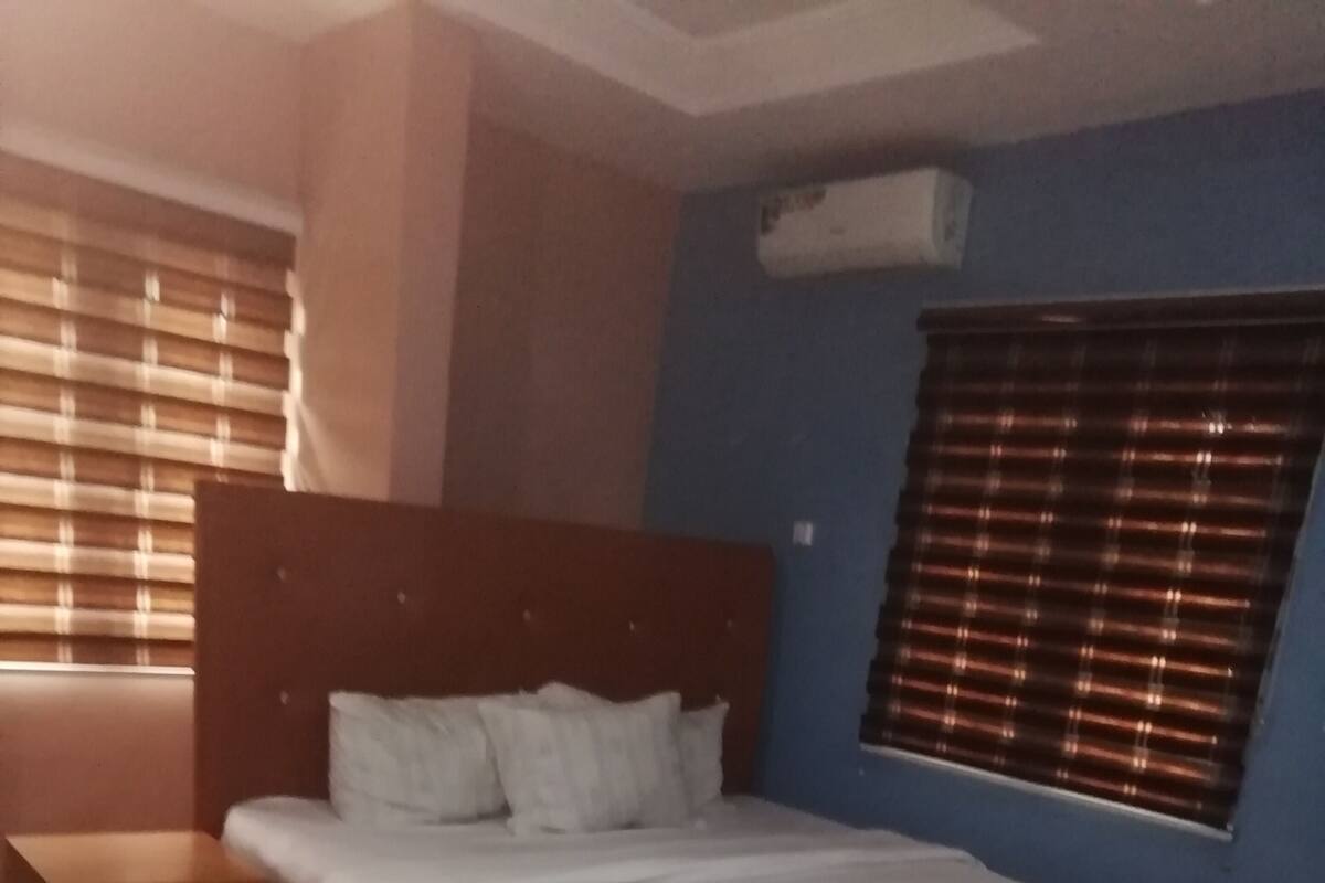 Een slaapkamer met een bed, een houten nachtkastje, een raam met zonwering en een airconditioning op de muur.