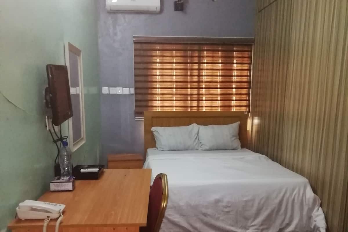 Een hotelkamer met een bed, een bureau met telefoon, een airconditioner en een raam met jaloezieën.