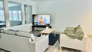 Ein modernes Wohnzimmer mit einem grauen Sofa, einem weißen Sessel, einem Couchtisch und einem Flachbildfernseher auf einem Standfuß.