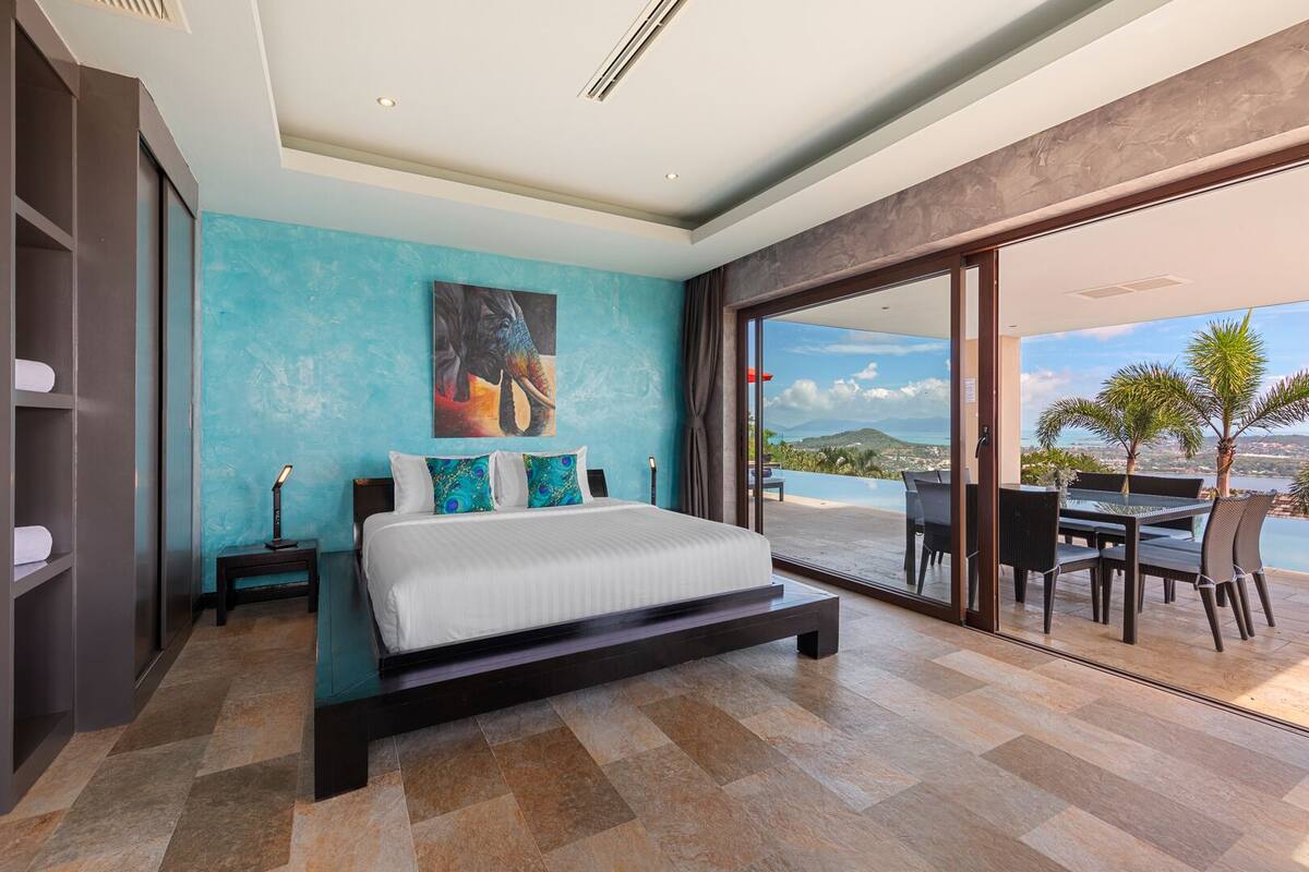 Une chambre moderne avec un grand lit, une vue sur la mer et un balcon aménagé avec du mobilier d’extérieur.