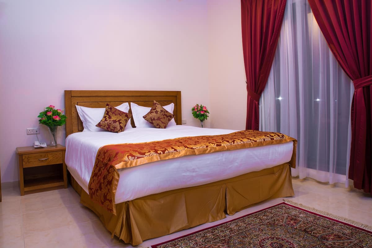 A hotel room with a large bed, wooden headboard, two bedside tables with lamps, a vase with flowers, and a view of the outside through sheer curtains.