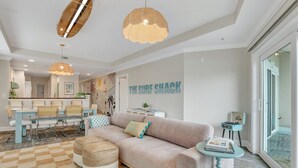 Een moderne woonkamer met een beige bank, een eethoek met een geruit kleed, en een muur met de tekst 'THE SURF SHACK'.