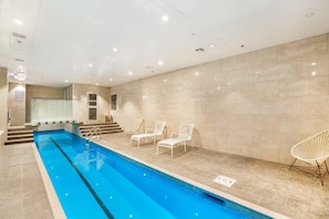 Une piscine intérieure à l’eau d’un bleu cristallin, entourée de murs recouverts de carreaux beiges et de chaises longues blanches.