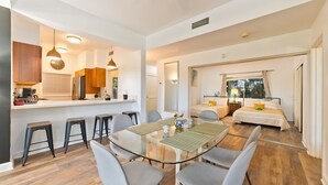 Un appartement moderne doté d’une table à manger en verre, de chaises grises, d’une cuisine avec des meubles en bois et d’une chambre comprenant un lit et une fenêtre.