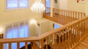 A wooden staircase with a chandelier, a dining area with tables and chairs, and patterned curtains.