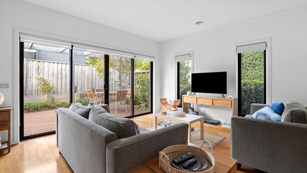 A modern living room with a grey sofa, a coffee table, and a TV on a wooden stand. There is a sliding glass door leading to an outdoor patio with a wooden fence and chairs. - Spring Central 29 Centrally Positioned 3 bedroom unit close to shops and beach (Torquay)