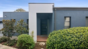 A modern two-story house with a grey exterior, a wooden front door, and a small porch area with a potted plant and a hedge. - Spring Central 29 Centrally Positioned 3 bedroom unit close to shops and beach (Torquay)