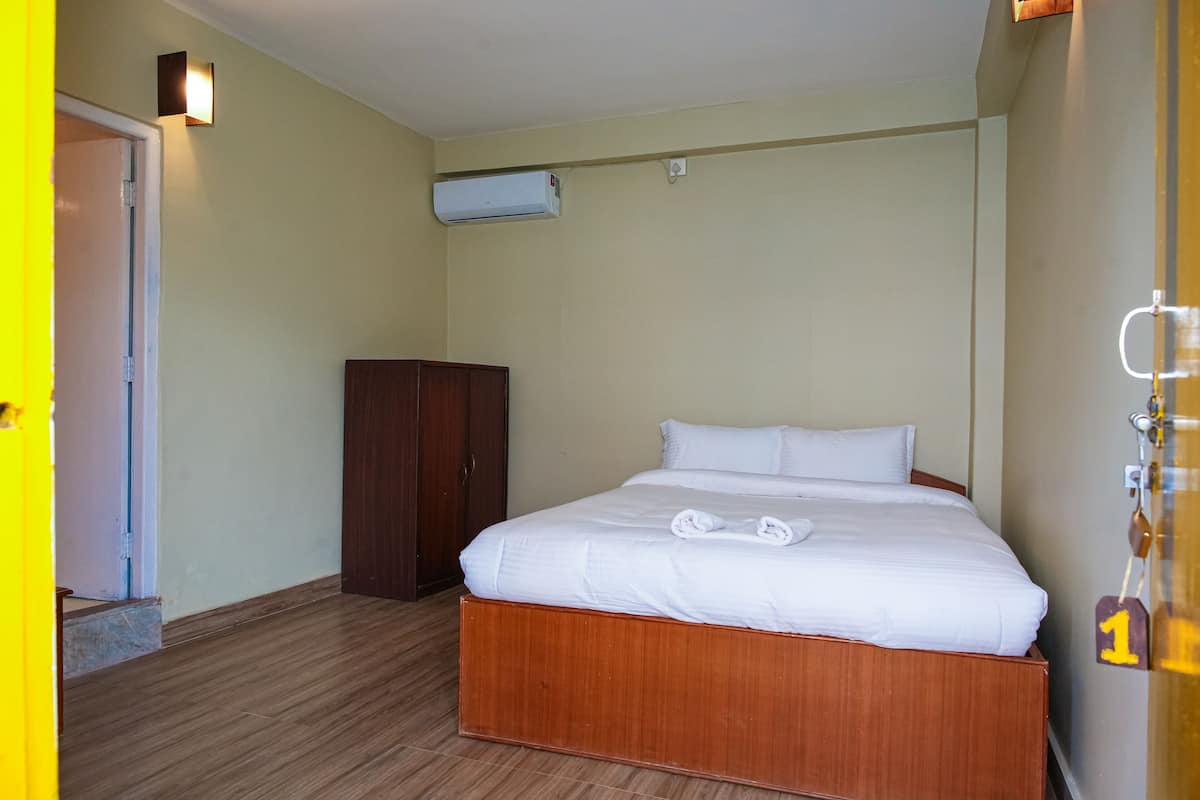 A hotel room with a wooden bed, white linens, a wardrobe, and an air conditioning unit.