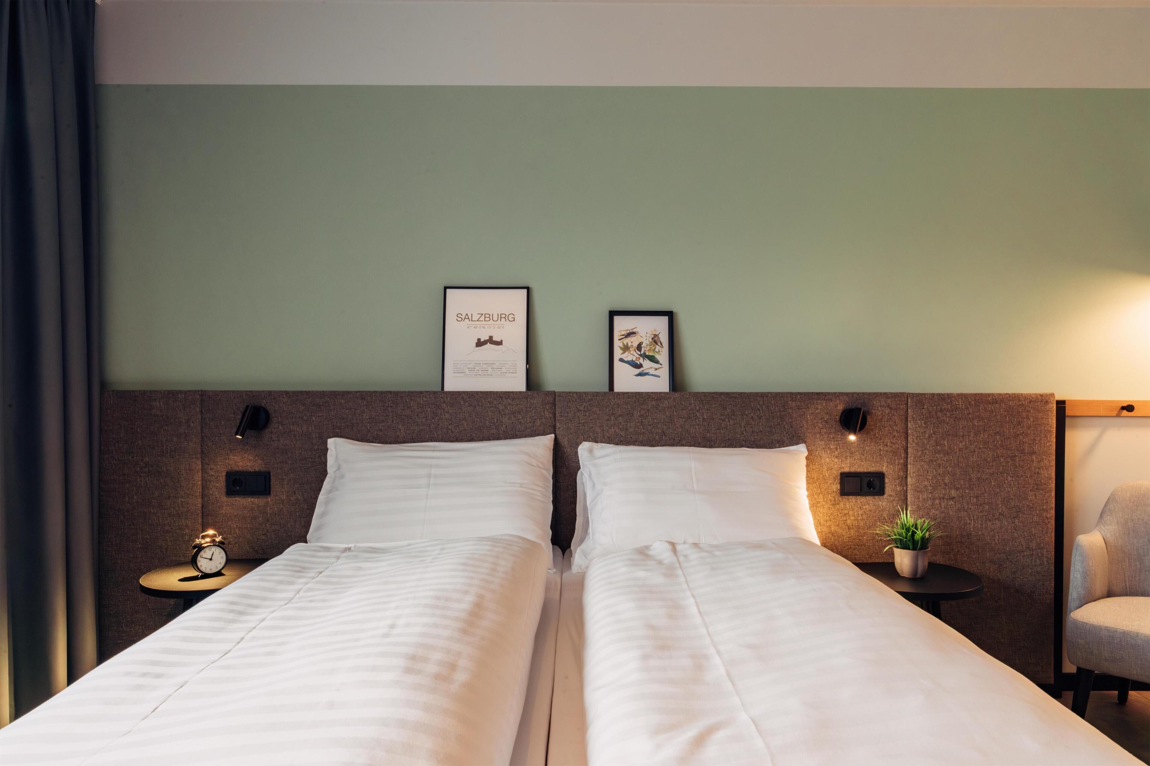A hotel room with two beds, a nightstand with a clock, a small table with a plant, and framed pictures on the wall.