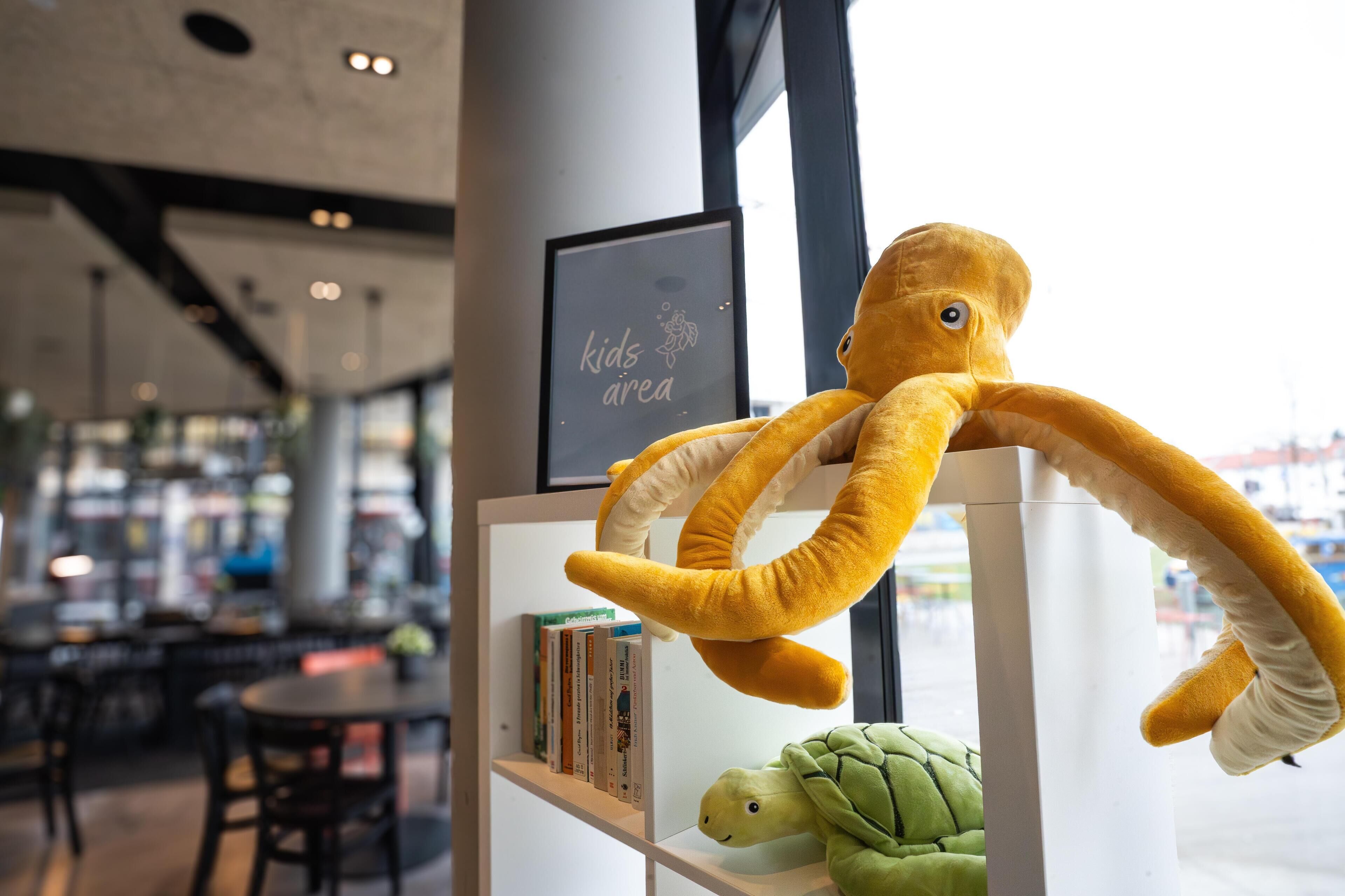 Un jouet en peluche représentant une pieuvre est suspendu à une étagère où se trouvent des livres, et un autre jouet, cette fois-ci une tortue, est placé en dessous.