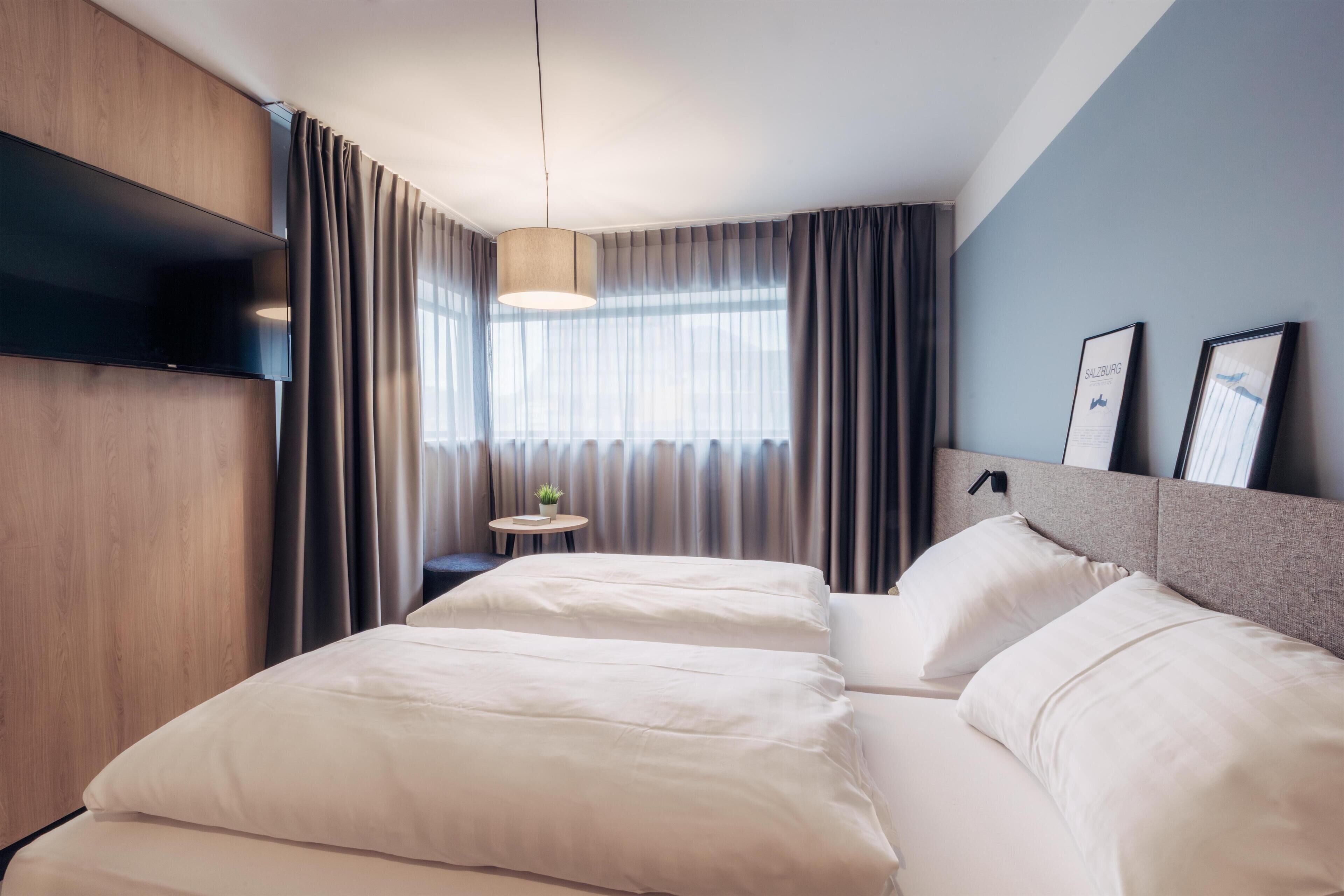 Ett modernt hotellrum med en stor säng, en platt-TV som är monterad på väggen, ett litet runt bord med en växt och ett stort fönster med gardiner.