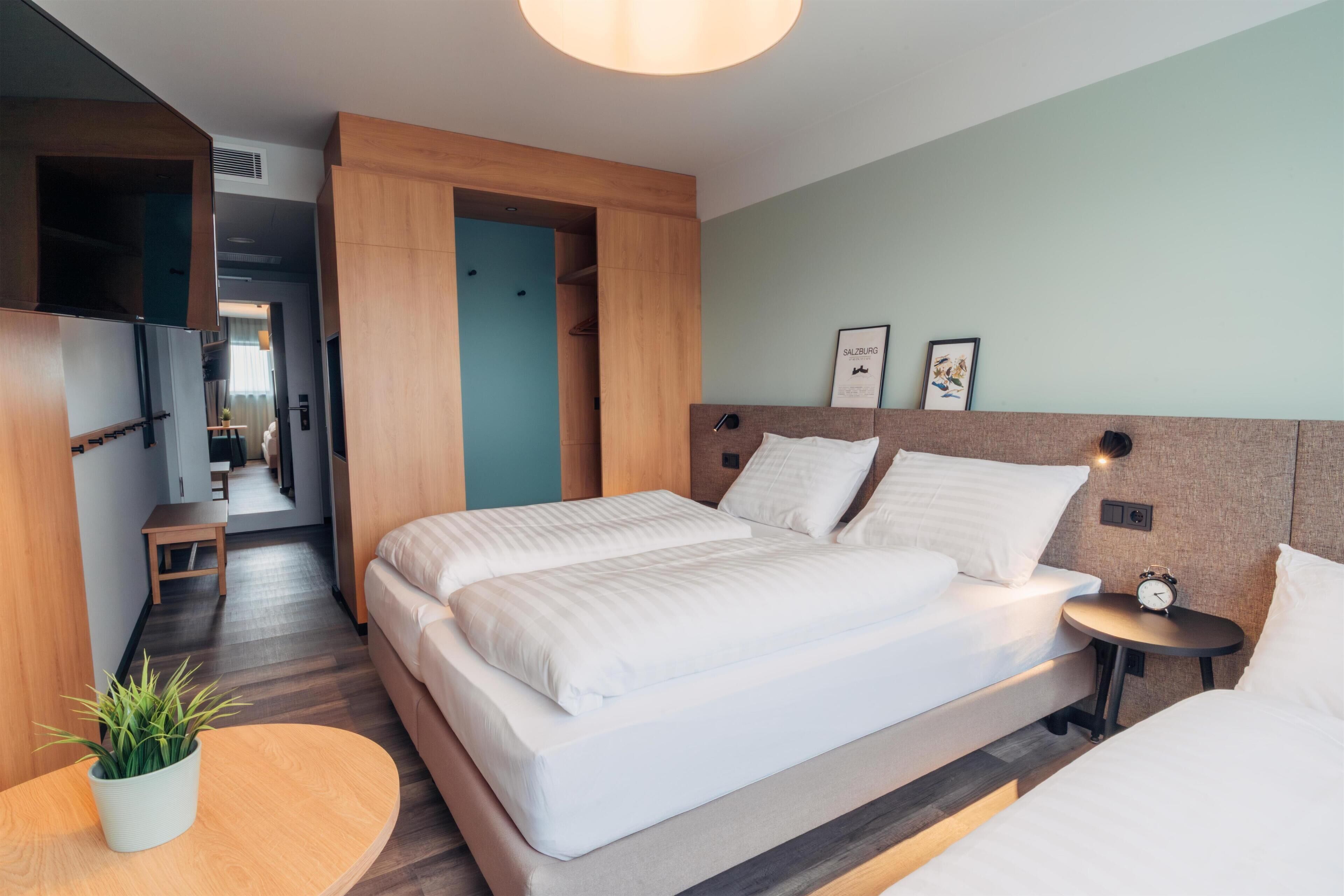 A modern hotel room with a large bed, bedside tables, a small seating area, and a TV mounted on the wall.