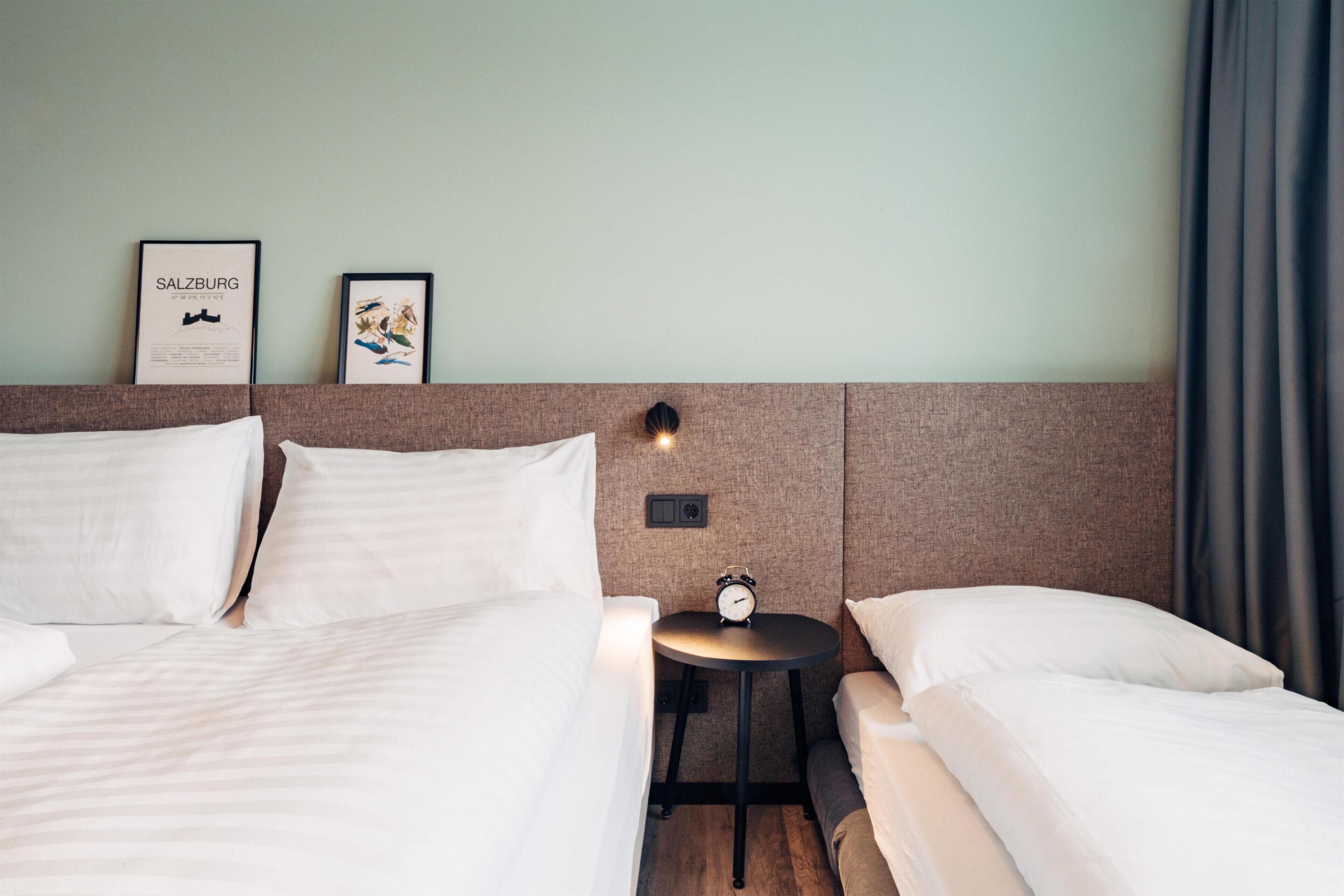 Ein Hotelzimmer mit zwei Betten, einem kleinen Tisch mit einer Uhr und zwei gerahmten Bildern an der Wand.