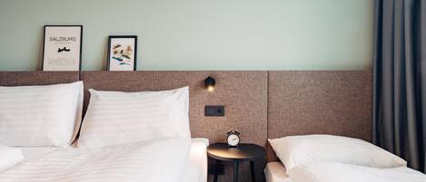 A hotel room with two beds, a small table with a clock, and two framed pictures on the wall.