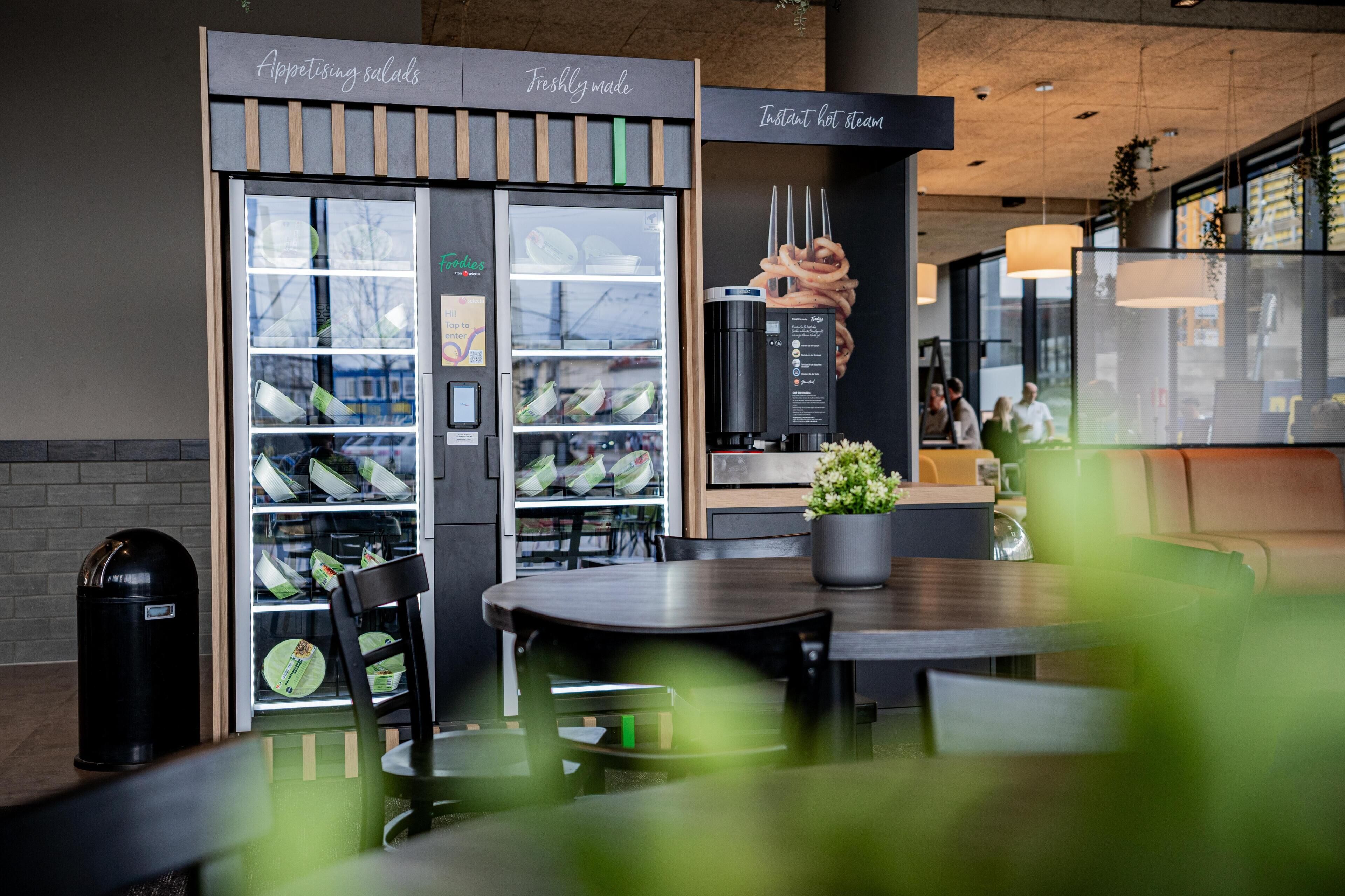 A restaurant with a glass display case filled with food items, a coffee machine, and a seating area with tables and chairs.