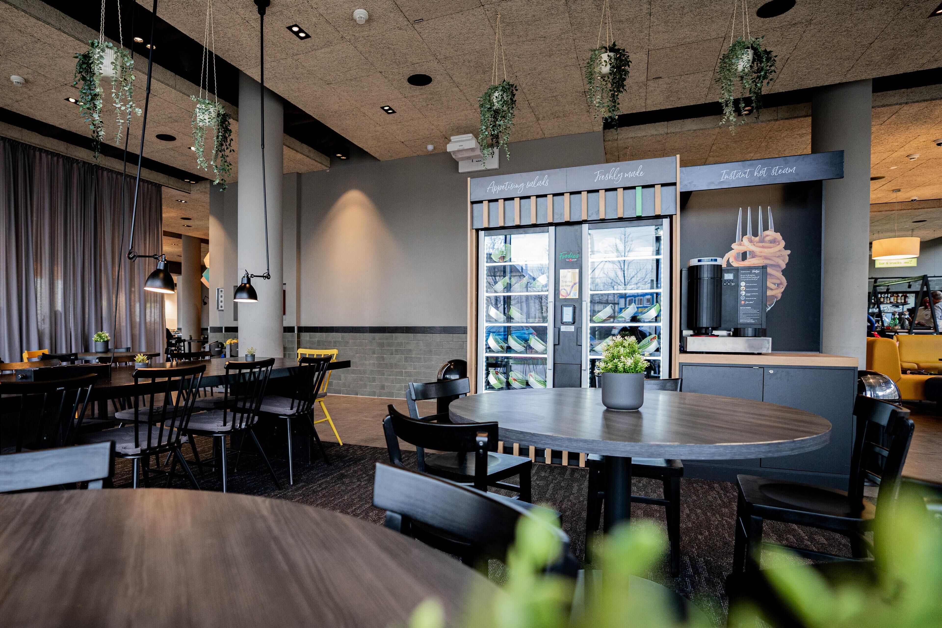 A modern restaurant with a central dining area featuring a round table and chairs, hanging plants, and a counter with a menu display.
