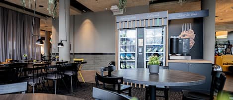 A modern restaurant with a central dining area featuring a round table and chairs, hanging plants, and a counter with a menu display.