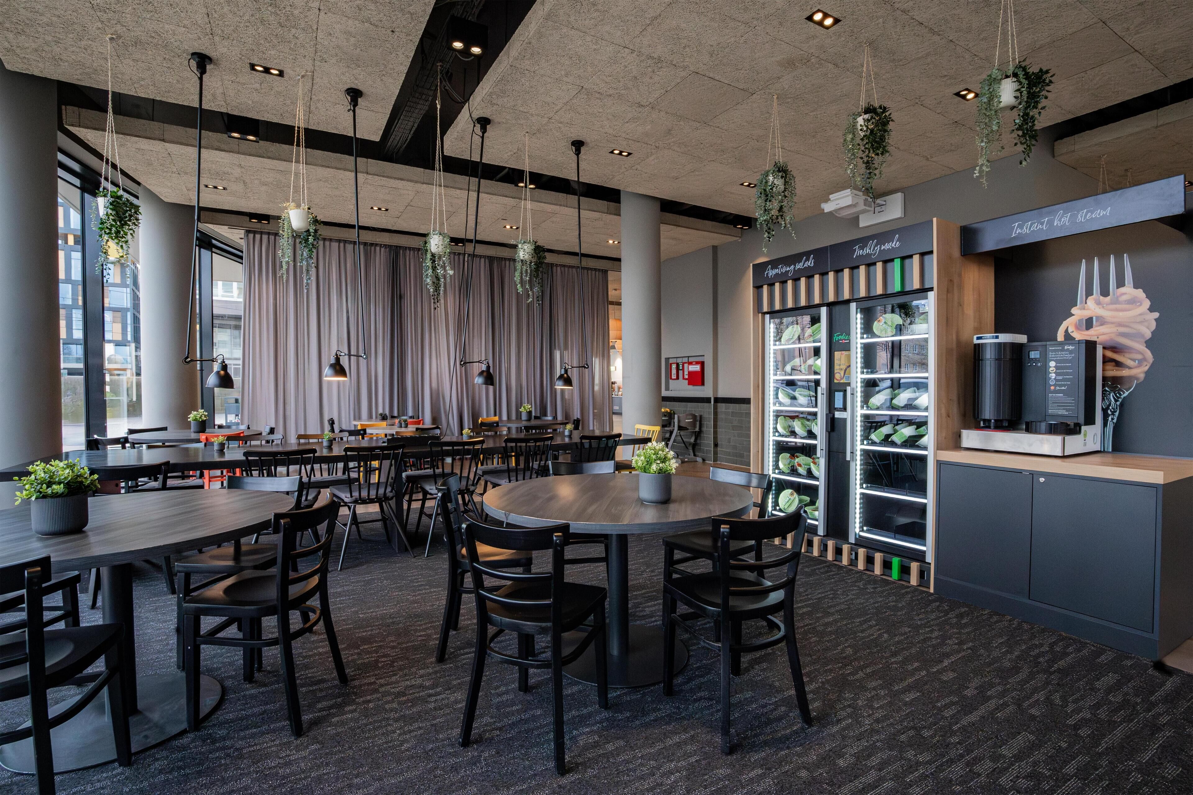 A modern dining area with round tables and chairs, hanging plants, and a coffee station.
