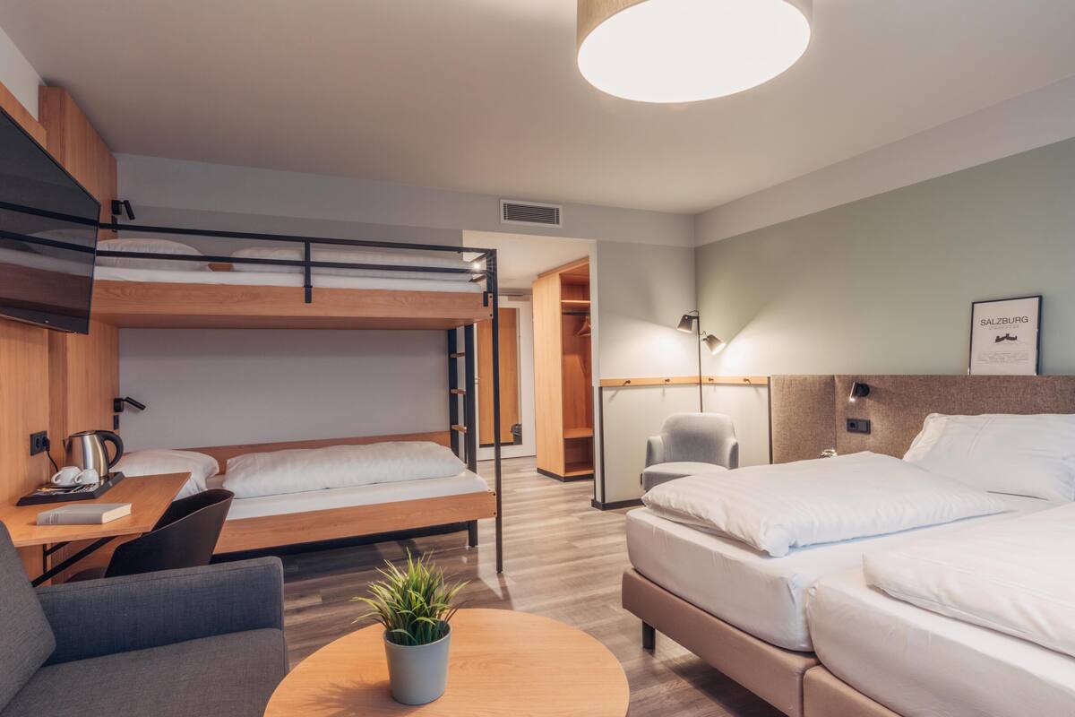 A modern hotel room with bunk beds, a desk with a laptop, a chair, a bedside table with a lamp, and a potted plant.